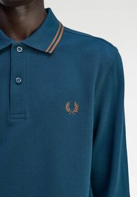 Polo shirt in teal scuro in tessuto texturizzato, con colletto classico con strisce marroni e logo ricamato sul lato sinistro del petto.