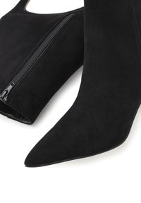 Stivaletti alla caviglia in suede nero con punta affusolata, dotati di zip laterale e una texture liscia. Il design è elegante e aderente.