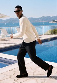 Jeune homme portant des lunettes de soleil, un pull blanc et un pantalon noir marchant à côté d'une piscine avec la mer et les montagnes en arrière-plan.