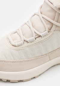 Beige hiking boot featuring a textured upper, padded tongue, and intricate lacing system. Rubber sole with traction pattern for grip.