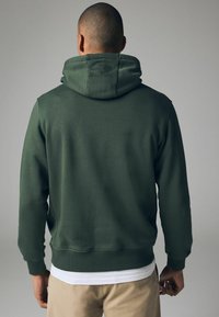 Homme aux cheveux courts portant un sweat à capuche vert foncé, un pantalon beige et une chemise blanche, dos tourné, sur un fond gris uni.
