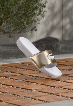 Biało-złota sandałka na płaskiej podeszwie z logo krokodyla, stojąca na drewnianej płytce na zewnątrz, w pobliżu zieleni i betonowej ściany.