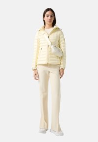 Yellow puffer jacket with a hood, zip closure, and side pockets, paired with cream trousers and a small white crossbody bag.