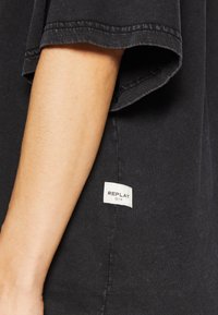 Camiseta de algodón negra con un corte relajado, que presenta una etiqueta de logo visible en la costura lateral y mangas ligeramente dobladas.
