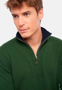 Pull zippé vert foncé avec un col bleu marine, présentant une fermeture éclair argentée et un tissu tricoté texturé. Gros plan sur les détails du vêtement.