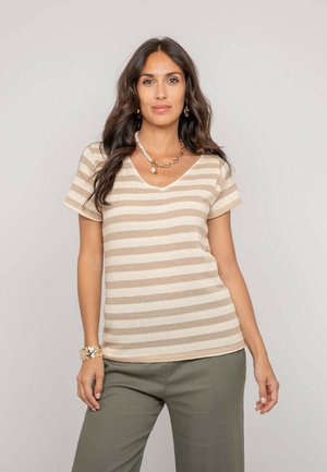 Femme aux longs cheveux foncés portant un haut à manches courtes beige et blanc rayé, un pantalon vert olive, un bracelet en or et des colliers superposés.