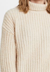 Beige Strickpullover mit lockerer Passform und hohem, geripptem Rollkragen. Hergestellt aus einem weichen, strukturierten Stoff mit sichtbaren Nähten.