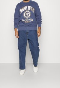 Felpa blu navy con grafica "Tommy Jeans", abbinata a jeans blu larghi e sneakers bianche. Vestibilità casual con polsini e orlo a costine.