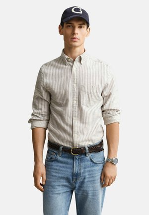 Junger Mann, der ein gemustertes Hemd mit hochgekrempelten Ärmeln, hellblaue Jeans, einen geflochtenen Gürtel, eine Armbanduhr und eine marineblaue Kappe mit weißem "G" trägt.