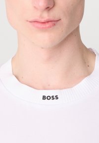 Bílé tričko s kulatým výstřihem vyrobené z hladké látky, s lemem na výstřihu a názvem značky "BOSS" v černé barvě na límci.