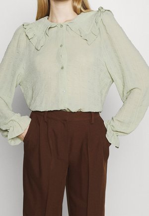 Lichtgroene blouse met ruches bij de kraag en mouwen, gemaakt van een gestructureerde stof en met een knoopsluiting aan de voorkant, gecombineerd met bruine hooguitlopende broeken.