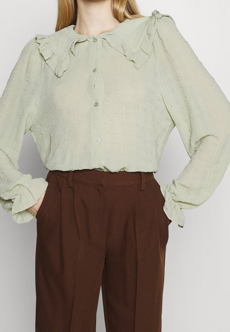 Light green blouse with ruffled collar and cuffs, featuring a textured fabric and button front, paired with brown high-waisted trousers.
