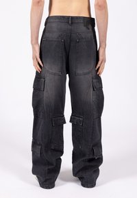 Osoba ubrana w luźne czarne spodnie cargo z wieloma klapkami kieszeni, widziana od tyłu na jednolitym białym tle.