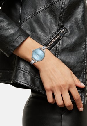 Montre en argent avec un cadran rond bleu clair, orné de pierres scintillantes, et un bracelet en cuir gris. Portée sur la manche d'une veste en cuir noir.