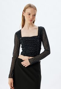 Schwarzes Crop-Top aus transparenter Stoff, mit langen Ärmeln, quadratischem Ausschnitt, Rüschendetails und weißen Punkten.
