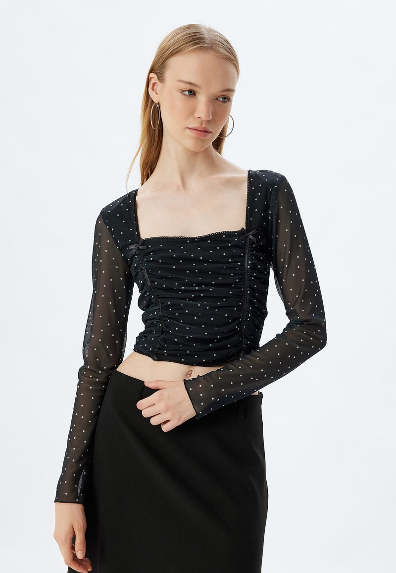 Schwarzes Crop-Top aus transparenter Stoff, mit langen Ärmeln, quadratischem Ausschnitt, Rüschendetails und weißen Punkten.