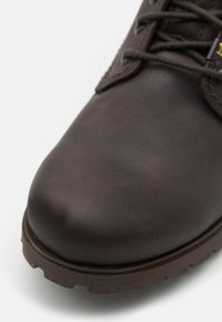 Bota de cuero marrón con puntera redondeada, superficie texturizada y suela de goma con profundas ranuras para mayor tracción. Se ven los cordones y los ojales de metal.