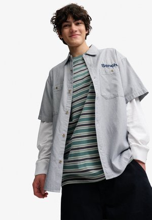 Ung mand med krøllet hår iført en grønstribet t-shirt under en grå kortærmet skjorte med "Bench." logo og sorte bukser.