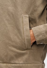 Χέρι που έχει μπει στην πλαϊνή τσέπη ενός μπεζ σακακιού με εμφανείς ραφές και μανσέτα με κουμπιά στο μανίκι.