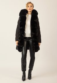 Schwarze Daunenjacke mit einem Kragen aus Kunstfell, die ein gestepptes Design, einen Reißverschluss und dazu passende schwarze, figurbetonte Lederhosen und Stiefel hat.