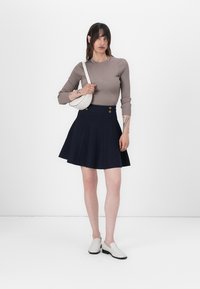 Beige geribbelde top met lange mouwen gecombineerd met een marineblauwe geplooide rok met twee gouden knopen. G gedragen met witte instapschoenen en een kleine witte tas.