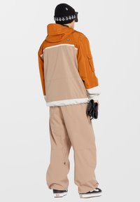 Orange och beige tvåfärgad jacka med huva, vita detaljer och en stor ficka. Beiga byxor med en avslappnad passform. Svart mössa med ett mönster.