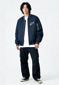 Giubbotto bomber blu navy con spilla di sicurezza e ricamo a forma di cuore, abbinato a pantaloni cargo blu navy larghi e sneakers nere. Il modello è in piedi contro uno sfondo chiaro.