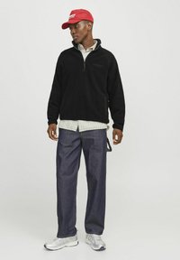 Sort fleece pullover med lynlås ved halsen, båret over en lysternede skjorte, parret med løse denimbukser og sølvfarvede sportssko. Rød kasket på hovedet.