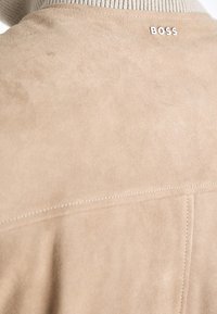 Gros plan de l'arrière d'une veste beige en daim avec le logo blanc BOSS près du col et des détails de couture visibles.