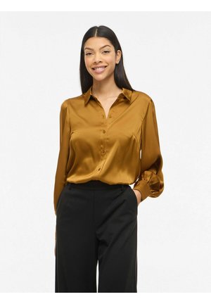 Tânără cu păr lung și închis la culoare, purtând o bluză din satin culoarea muștarului și pantaloni negri, zâmbind cu o mână în buzunar.