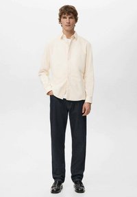 Camicia beige chiaro con bottoni in tessuto morbido, abbinata a pantaloni di denim scuro e mocassini neri, per un outfit casual.