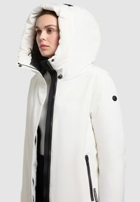 Hvid polstret jakke med en stor hætte, udstyret med en sort lynlås og detaljer, sidelommer og glat, blankt materiale.