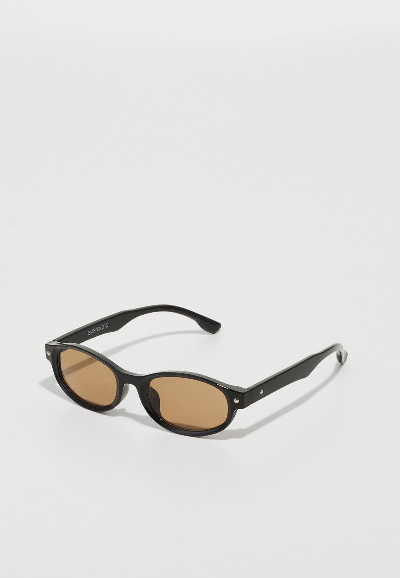 Black sunglasses with oval frames, brown tinted lenses, and silver accent hardware on the frame. Smooth texture and sleek design.