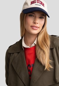 Casquette beige avec "BIG STAR JEANS" en rouge, trench-coat olive vert, sweat-shirt rouge et col rayé visible ; cheveux lisses, teint naturel.