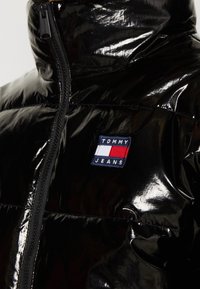 Czarna, błyszcząca kurtka puchowa z wysokim kołnierzem, z centralnym zamkiem błyskawicznym oraz naszywką z logo "TOMMY JEANS" w niebieskim, czerwonym i białym kolorze.