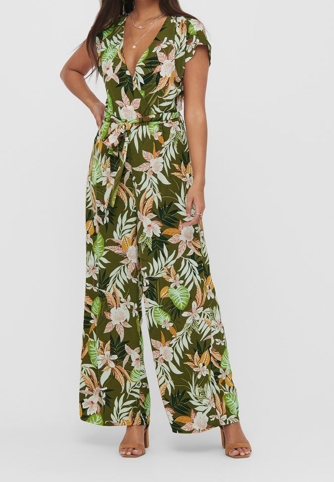 Femme portant une combinaison pantalon verte à jambes larges avec imprimé floral, manches courtes, col en V profond et ceinture nouée à la taille, associée à des talons ouverts.