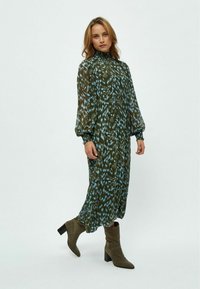 Langes grünes Kleid mit blauem abstraktem Muster, hohem Kragen und Ballonärmeln. Kombiniert mit olivfarbenen Stiefeletten und glatter Textur.