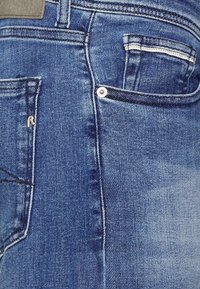 Donkerblauwe denim jeans met een achterzak, zilveren klinknagel en stikseldetails. Textuurstof met een lichte vervagingseffect.