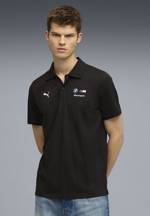 Schwarzes Polo-Shirt mit subtiler Textur, mit BMW- und Puma-Logos auf der Brust; kurze Ärmel und ein Kragen. Getragen mit hellblauen Jeans.