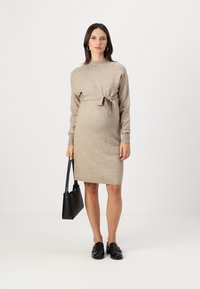 Abito lavorato a maglia beige con colletto alto e vita regolabile, maniche lunghe, polsini a coste, indossato con mocassini neri e una borsa a spalla nera.