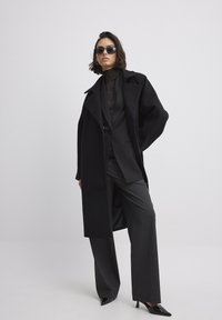 Abrigo largo negro sobre un traje negro a medida, combinado con pantalones de pierna ancha grises y tacones negros de punta. Los accesorios incluyen gafas de sol negras.