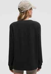 Chemise noire à manches longues avec une coupe décontractée et un col rond, fabriquée en tissu doux. Associée à une casquette beige, vue de dos.