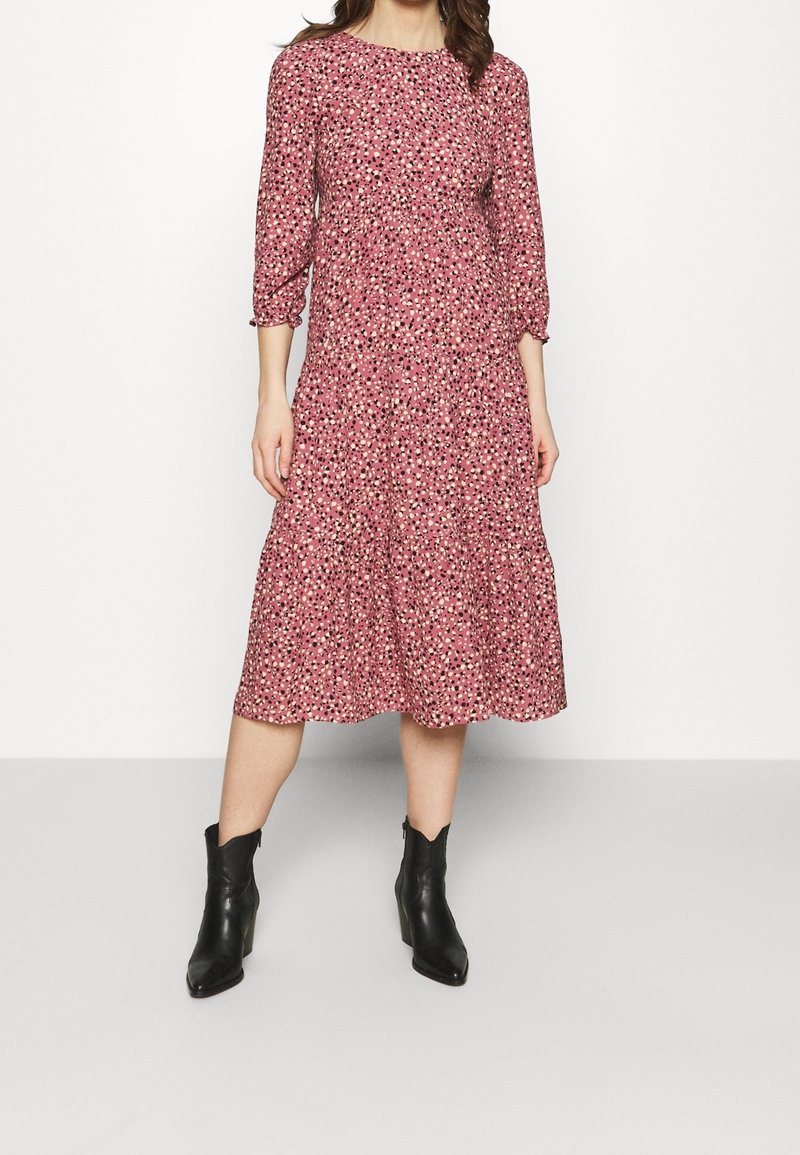 Femme portant une robe midi rose à petit motif floral et manches trois quarts, associée à des bottines noires.