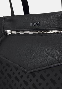 Sac fourre-tout noir avec une finition texturée, comportant une poche zippée et un logo en relief. Le design inclut un motif géométrique et des accents lisses.