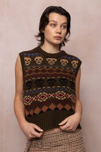 Jauns cilvēks ar īsiem tumšiem matiem valkā bez piedurknēm brūnu adītu džemperi ar skeleta motīviem un raibu bikses, stāvot pie bēša sienas.