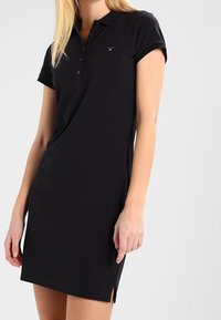 Robe polo noire avec un col, des manches courtes et une patte de boutonnage. Présente des fentes sur les côtés et un petit logo sur la poitrine.