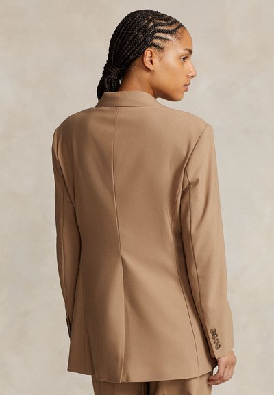Femme aux cheveux tressés portant un blazer camel sur mesure, vue de dos sur un fond neutre.