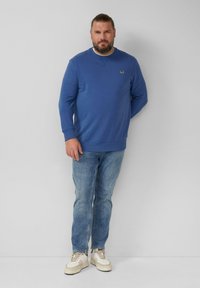 Felpa blu con polsini e orlo a costine, caratterizzata da un piccolo logo sul petto. Abbinata a jeans azzurri chiari e sneaker bianche.