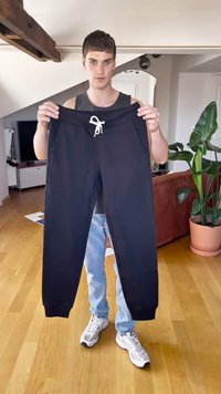 Pantaloni della tuta neri con cordino, vita a coste e tasche laterali, tenuti da una persona in un ambiente informale con pavimento in legno.