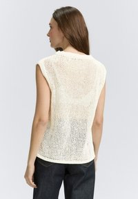 Strickoberteil in Creme ohne Ärmel mit offenem Muster, Rundhalsausschnitt und Rippkanten. Das Model trägt dunkelblaue Jeans mit Gesäßtaschen.